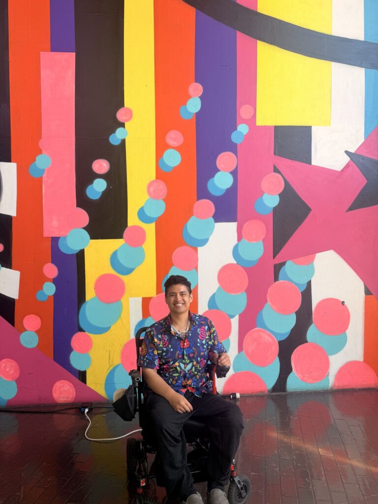 Mike smiling in his power wheelchair in front of a colorful geometric mural