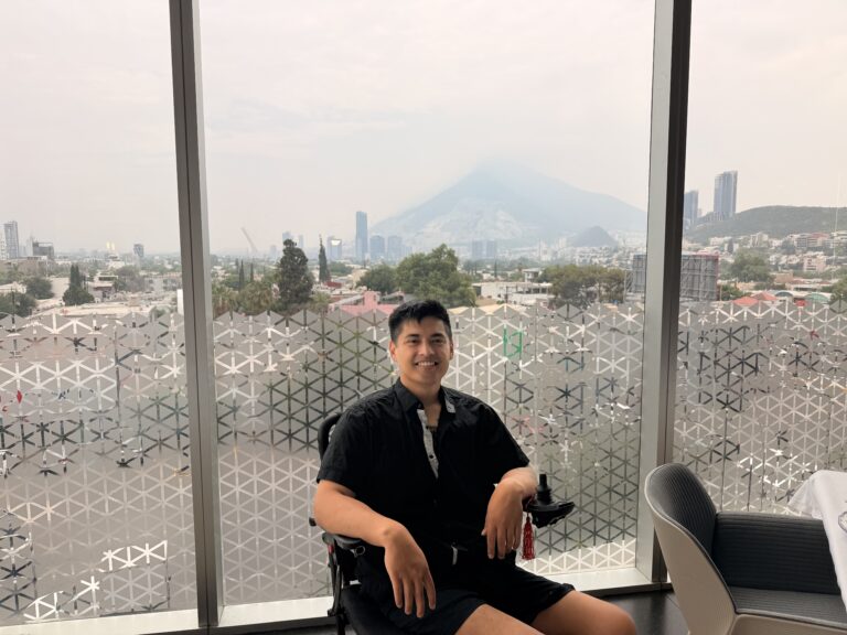 Miguel seated by a floor to ceiling window with a city skyline and mountain behind him