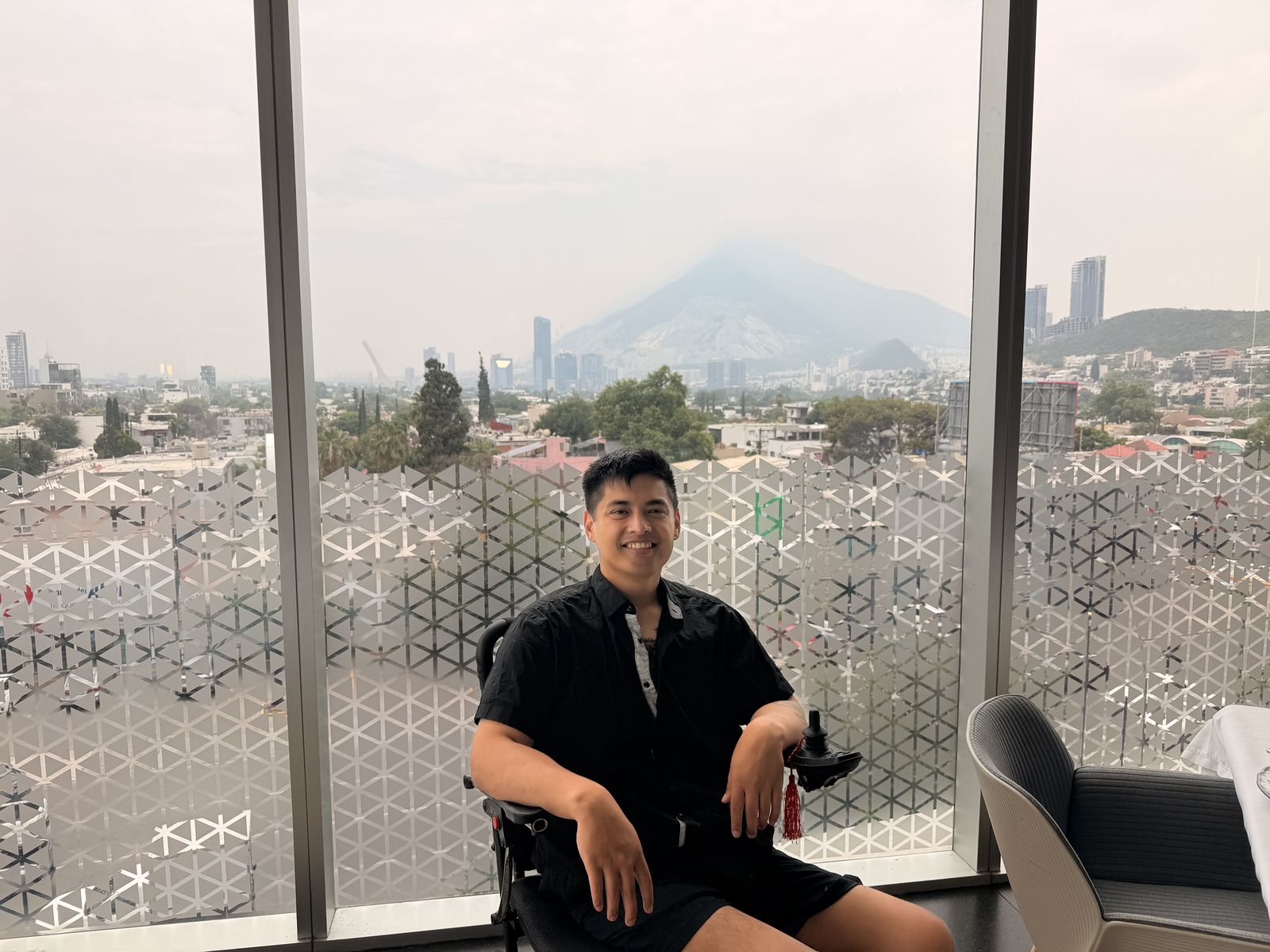 Mike seated by a floor to ceiling window with a city skyline and mountain behind him