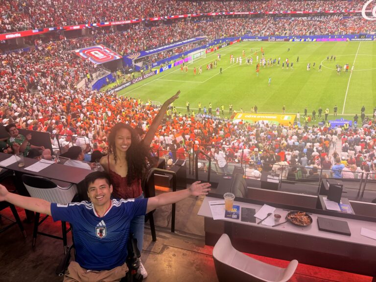 Cielo and Miguel cheering from an accessible suite at a packed soccer stadium