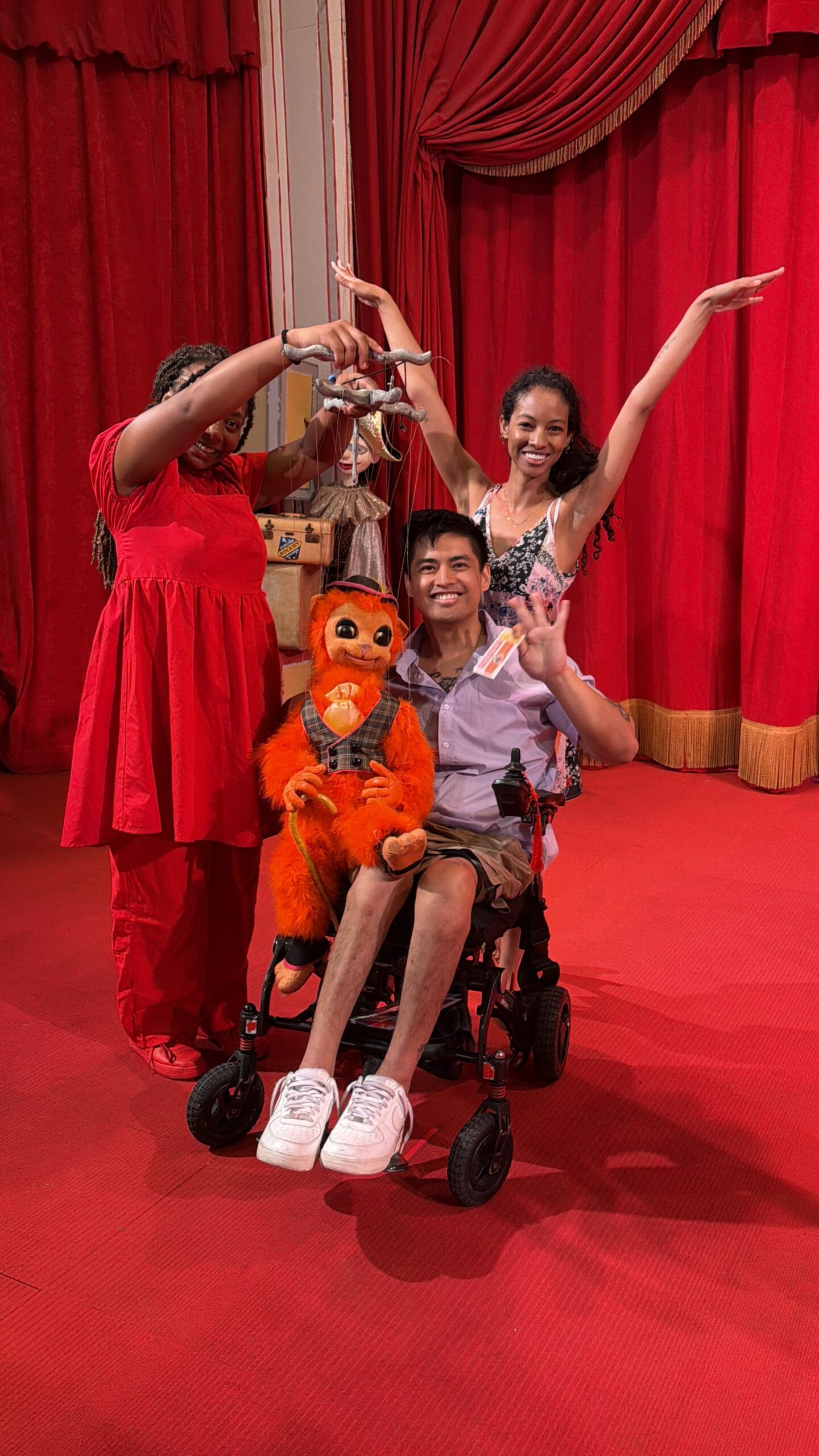 Mike holding an orange monkey puppet with two friends in front of red theater curtains