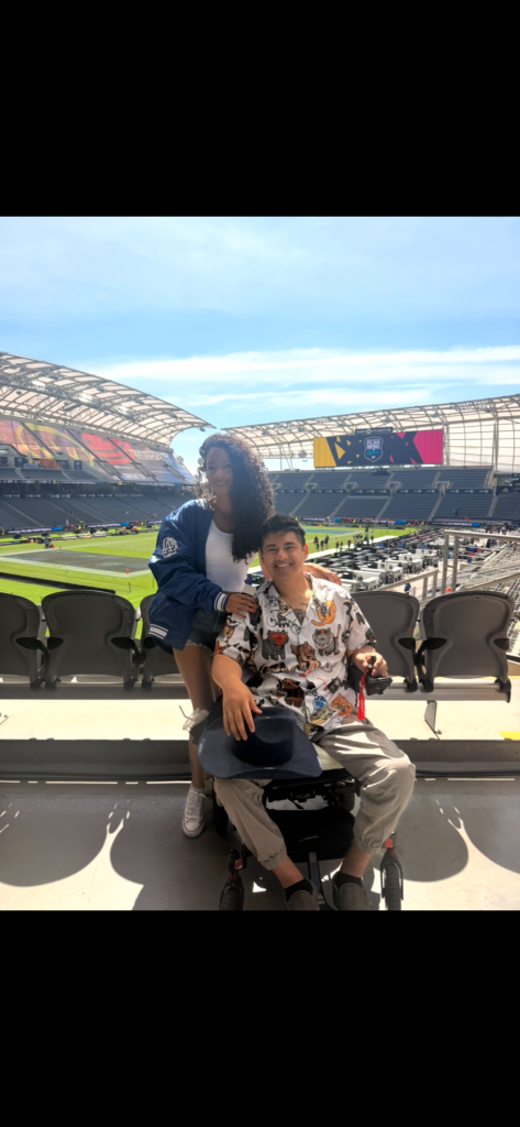 Cielo leaning on Miguel in accessible seating at an open air stadium