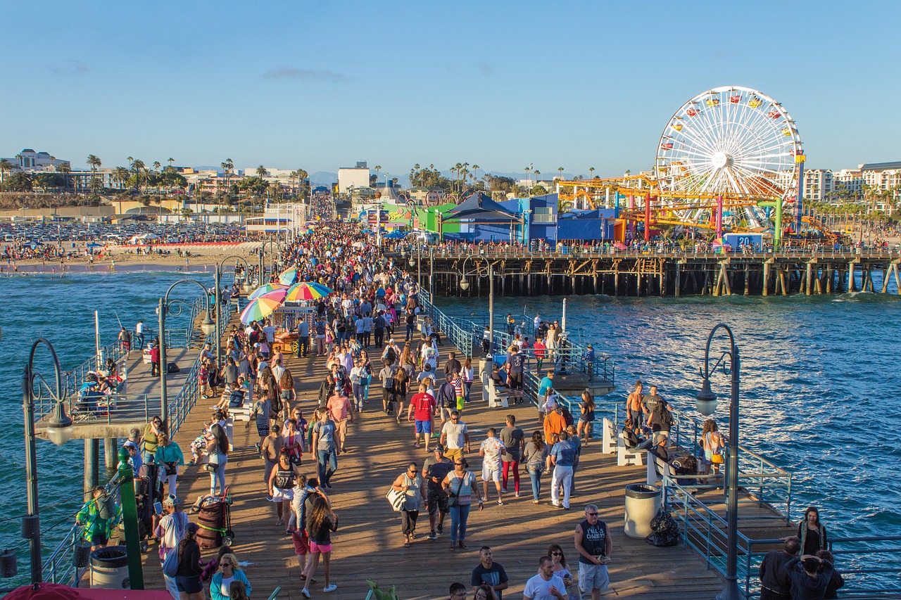 Crowds walking the wooden deck of Santa Monica Pier on a sunny afternoon, Ferris wheel behind them.