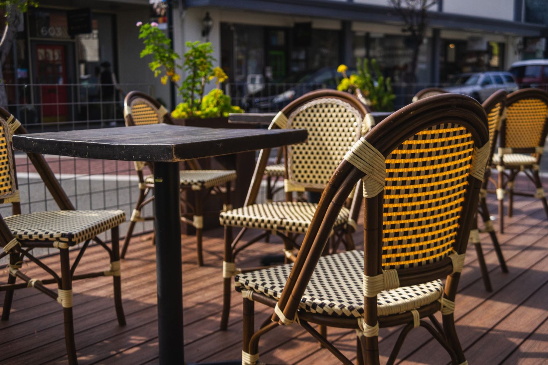 Cafe tables and woven bistro chairs on a raised wooden sidewalk dining deck beside a city street