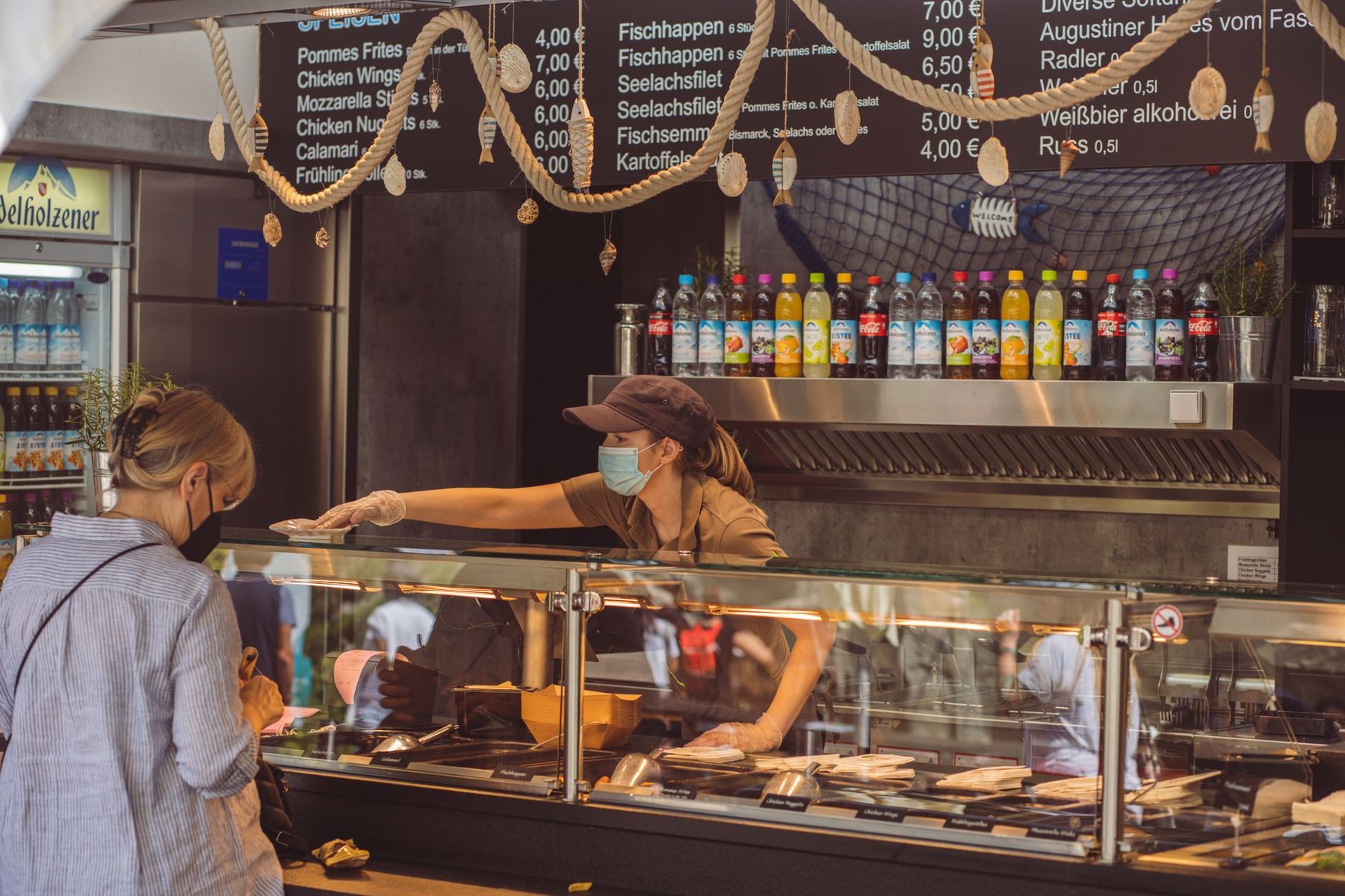 Customers ordering at a food stall with a tall glass display counter and a menu board mounted above it