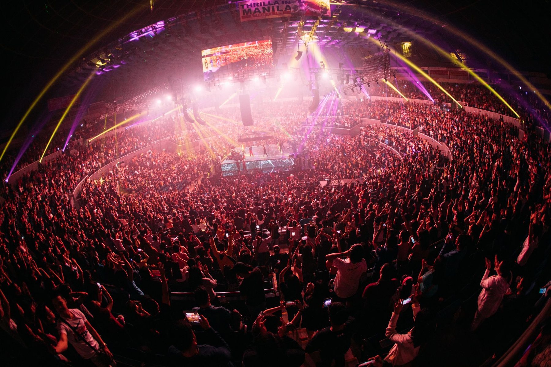 A packed arena crowd lit in pink and purple during a concert, seen from the upper level