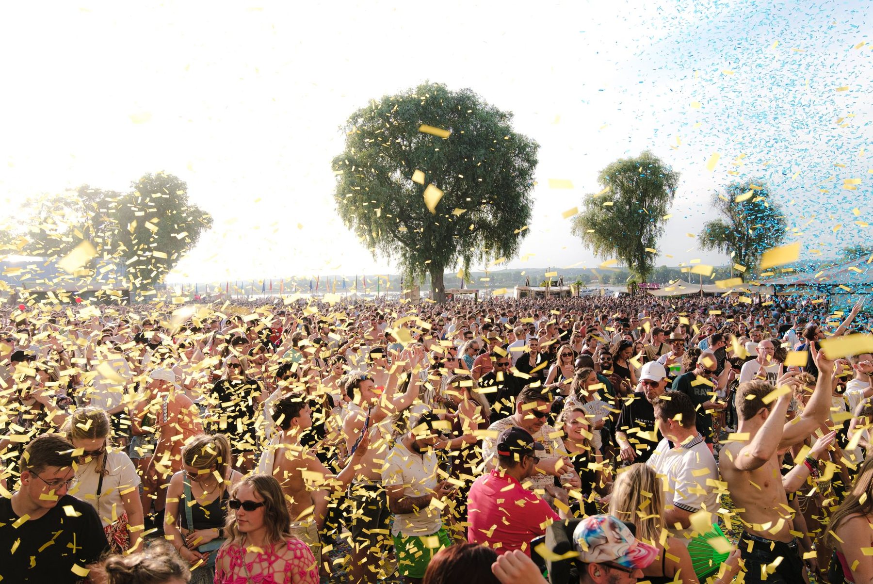 A festival crowd filling an open field beside a row of trees, with confetti falling over the audience