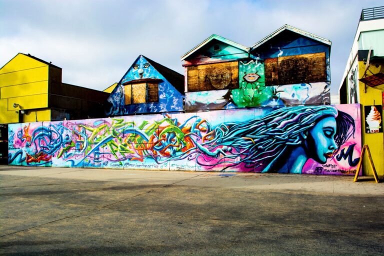 A long colorful mural painted along a street wall with an empty paved lot in front of it