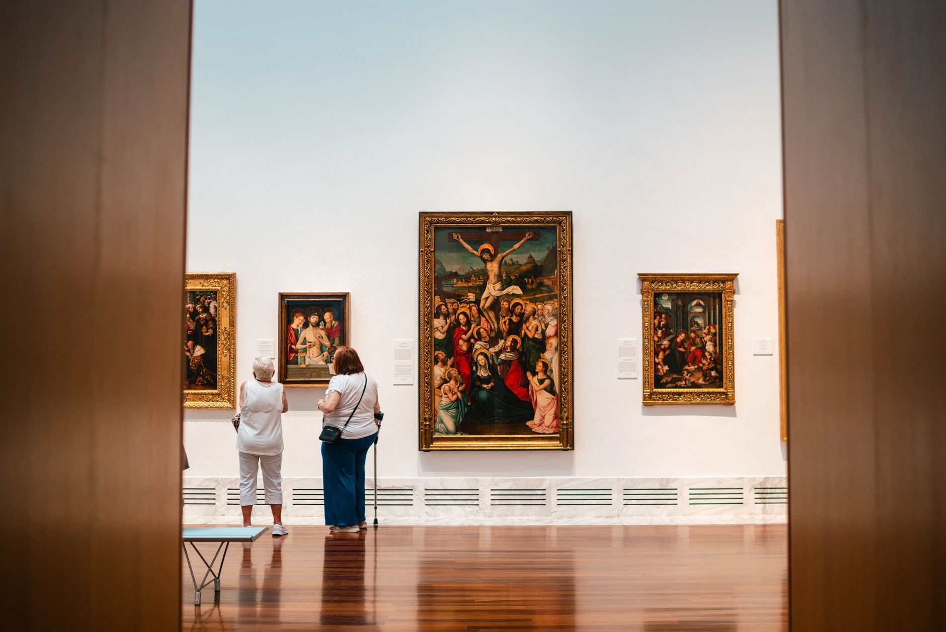 Two visitors standing in front of large framed paintings in a wide museum gallery with a polished wood floor