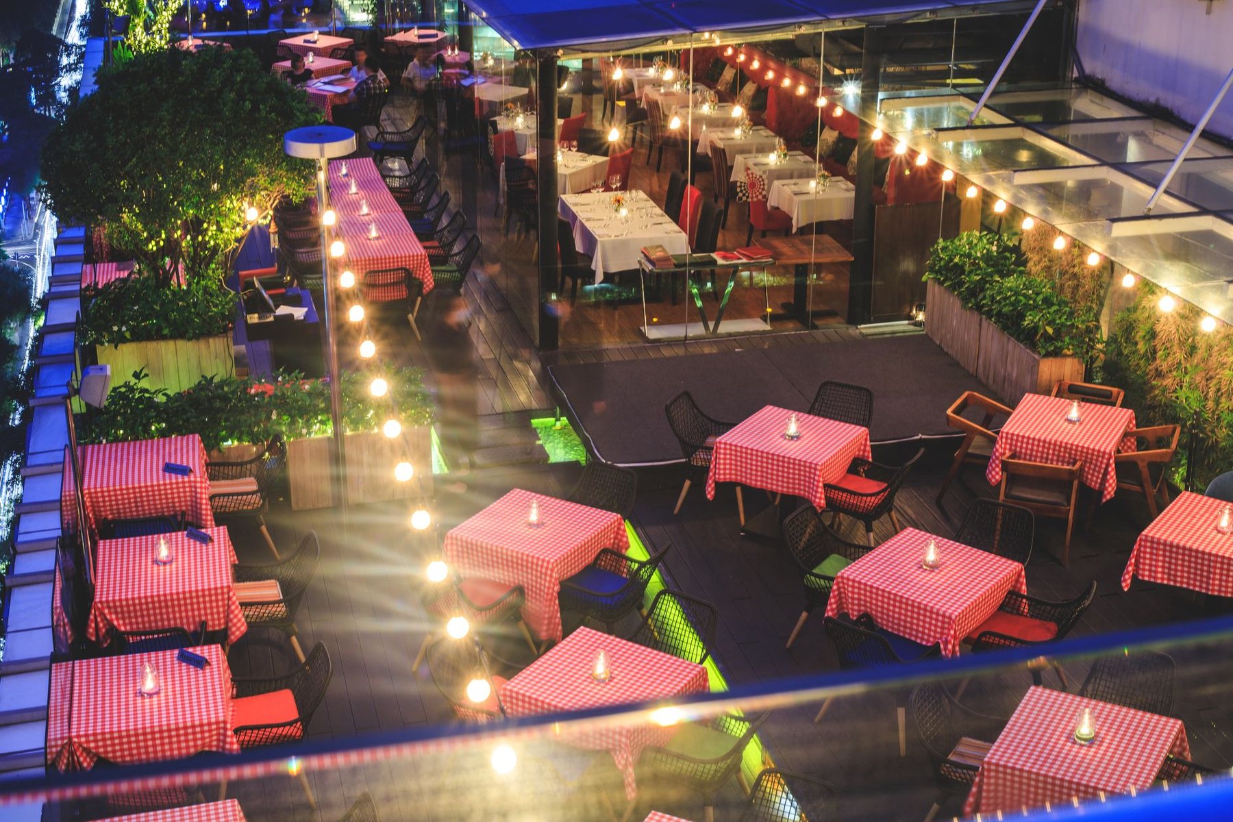 An outdoor terrace bar at night seen from above, with red-topped tables and strings of lights overhead