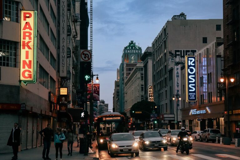 A busy street in downtown Los Angeles at dusk, with lit storefront signs, a parking sign and traffic in both directions
